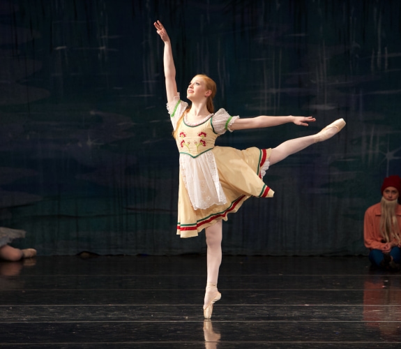 A student performing on a stage while wearing pointe shoes and a yellow costume
