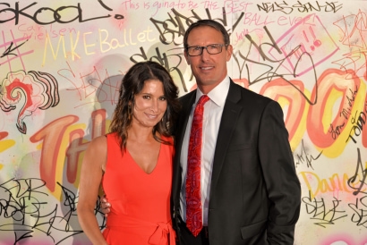 A couple smiling in formal attire, in front of a graffiti wall backdrop